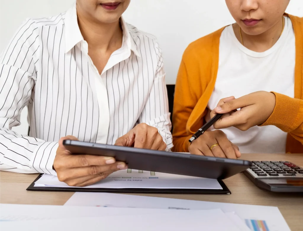 Two women sitting side by side pointing at a tablet with a clipboard and calculator before them indicating them calculating compensation for land acquisition.