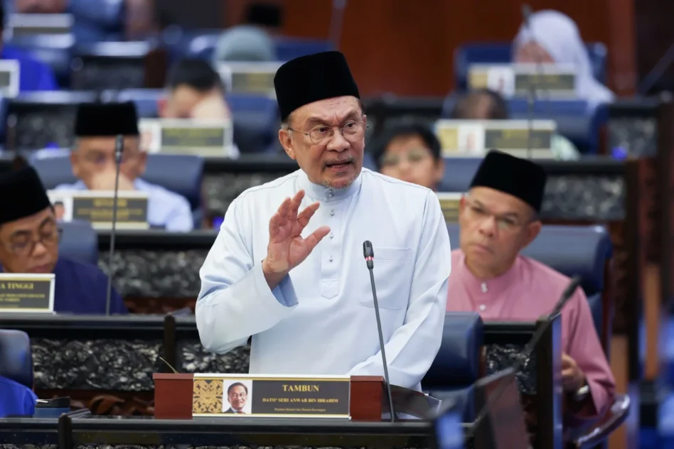 Budget 2026 Malaysia, Prime Minister and Finance Minister Dato’ Seri Anwar Ibrahim wearing a traditional Malay outfit — a white baju Melayu and black songkok — speaking passionately at a parliamentary session. He stands at a podium with a microphone, addressing the assembly, while several other members, dressed in formal attire, listen attentively in the background. The setting appears to be a formal government chamber with nameplates and seating rows.