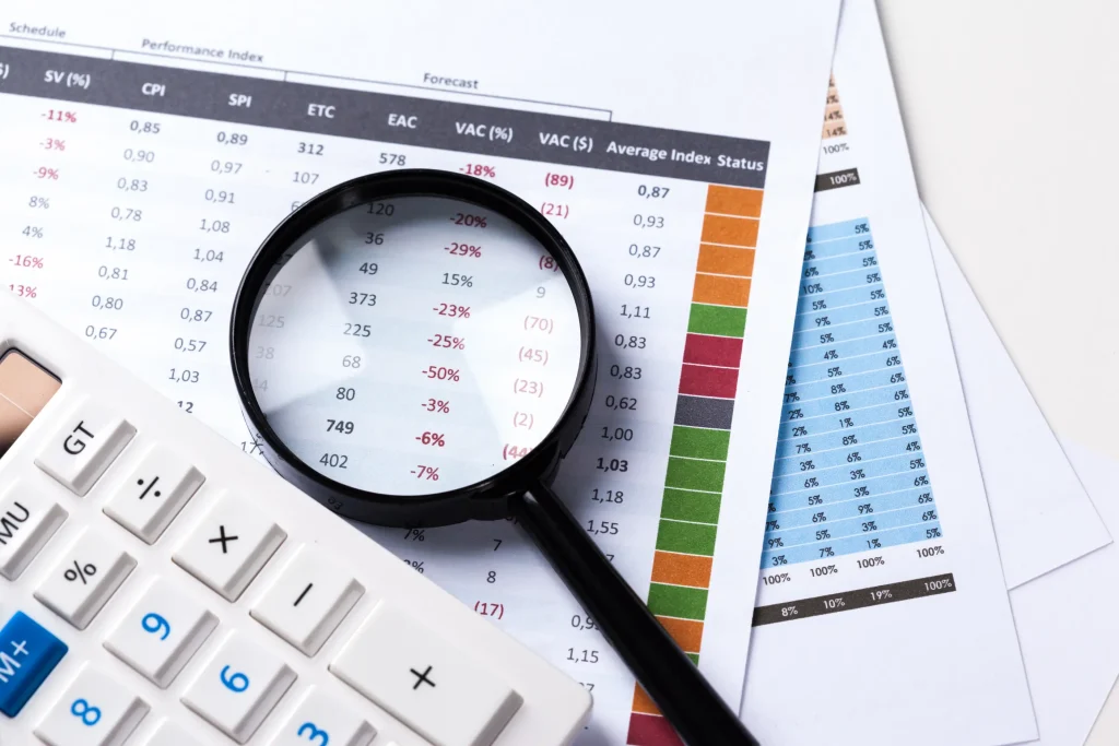 Budget 2026 Malaysia illustration. It shows shows a close-up view of financial documents on a desk. A magnifying glass lies on top of a printed spreadsheet, zooming in on a column of red and black numerical data. The sheet features performance and forecast figures, with some numbers showing negative percentages highlighted in red. Next to the papers is a white calculator, partially visible in the bottom left corner.