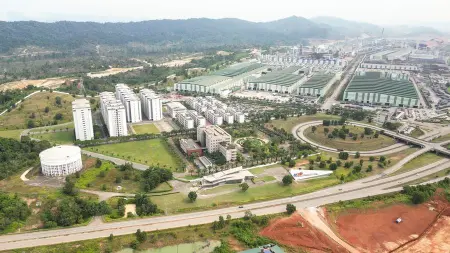 For the budget 2026 malaysia, this image shows an aerial view of an expansive industrial and residential area surrounded by greenery and hills. In the foreground, there are modern apartment buildings and neatly arranged roads, while the background features large factory or warehouse complexes with long green rooftops. The layout appears organized, with landscaped areas and open spaces separating the zones — reflecting industrial development and urban planning in progress.