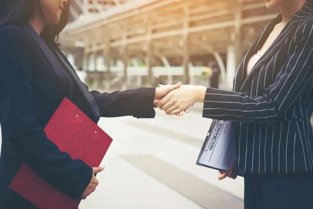 Two businesswomen in formal suits shaking hands outdoors, symbolising a successful corporate deal by a merger and acquisition company in Malaysia. One holds a red folder and the other a clipboard labeled “Insurance.”
