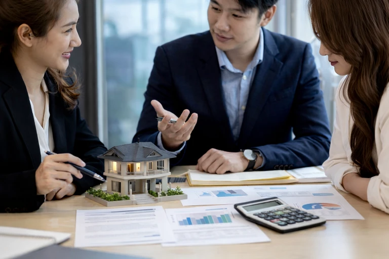 Housing loan rates consultation between three Asian professionals seated at a desk, discussing property financing with a detailed house model, financial charts, and a calculator on the table, while the woman on the right listens attentively with her hands tucked close to her body in a bright modern office setting.