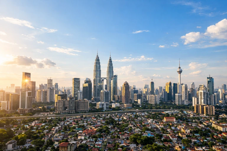 Real Estate Investment in Malaysia post with a bright aerial photograph of Kuala Lumpur’s city skyline under a clear blue daytime sky, featuring the iconic Petronas Twin Towers standing prominently at the center and the KL Tower visible to the right. In the foreground, a dense residential neighborhood of low-rise houses with red and brown rooftops stretches across the landscape, intersected by roads and greenery. Behind the residential area, modern high-rise office and commercial buildings form a dense urban core, reflecting sunlight. The overall scene shows a vibrant Malaysian city with a mix of residential communities and towering skyscrapers under bright natural daylight and scattered clouds.