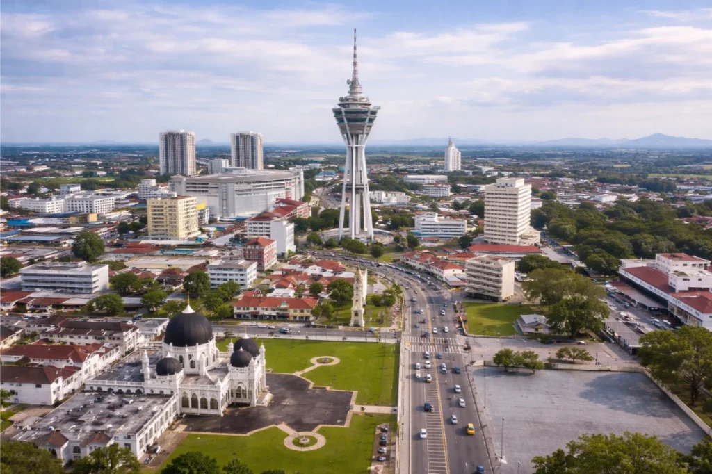 Aerial cityscape of Alor Setar, featuring the iconic Alor Setar Tower rising prominently at the center of a well-planned urban landscape. In the foreground, the Zahir Mosque with its distinctive black domes and white structure sits beside open green spaces and road networks with light traffic. Surrounding the area are mid-rise residential and commercial buildings, with a mix of traditional rooftops and modern developments extending toward the horizon. The image highlights the balance of heritage landmarks and growing urban infrastructure within Kedah’s evolving property market.