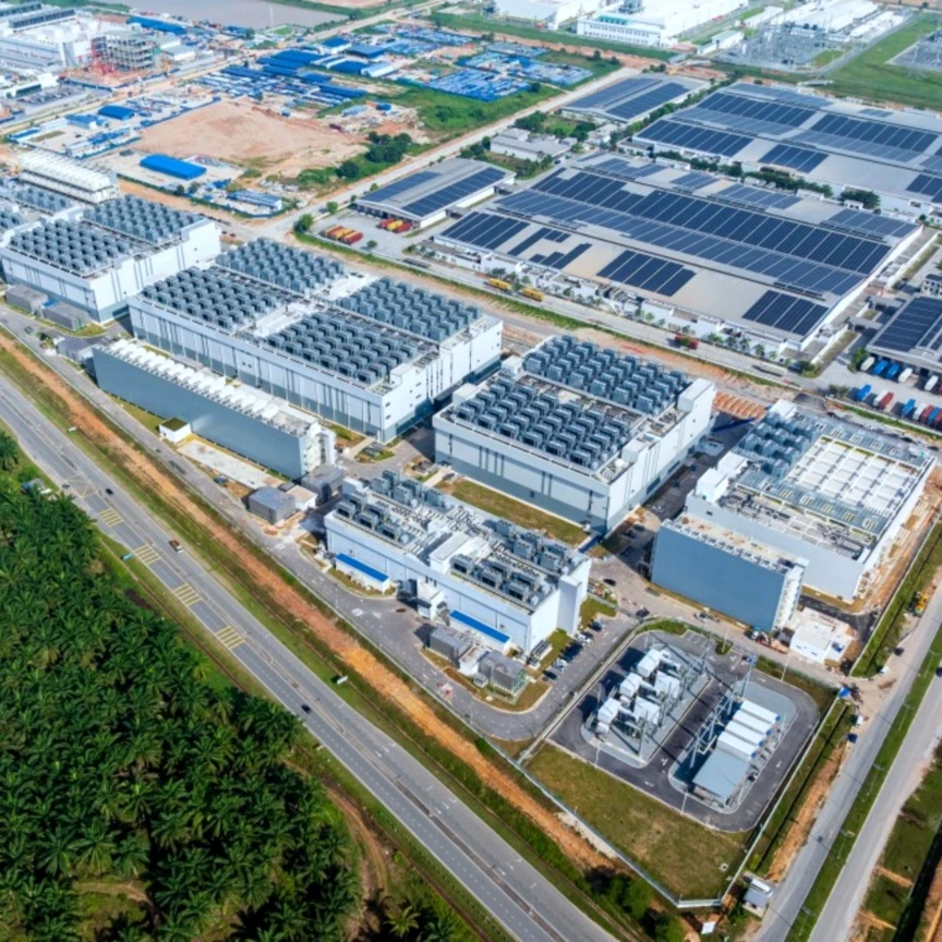 Aerial photograph of a large-scale industrial facility featuring multiple modern factory buildings with structured layouts and advanced infrastructure. Several rooftops are fitted with solar panels, indicating energy-efficient operations, while surrounding areas include access roads, parking zones, and additional industrial developments under construction. The facility is strategically located near main roads and greenery, showcasing the growth of high-tech industrial property and sustainable development within Kedah’s expanding industrial landscape.